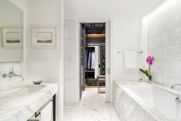 NYC EAST VILLAGE APARTMENT RENOVATION - An apartment combined from two smaller units in an industrial-converted-to-residential building in New York City’s East Village presented an opportunity to create a large master bathroom and dressing space, atypical for a Manhattan apartment.   

The well-traveled owner enlisted architect Adi Gershoni to design a space with the feel of his favorite luxury hotel. The highly detailed space and the choice of materials, including natural marble slabs and tile, concealed lighting and state-of-the-art plumbing fixtures, deliver this level of quality – and something more: As a custom, one-of-a-kind design, it retains an individuality and hominess that its inspiration lacks.   

The new plan joined what used to be a foyer in one of the previous apartments, a small bathroom and several separate closets into a combined space that flows off the master bedroom. In adapting this hospitality-inspired concept to a home, the challenge was to retain the luxe, minimalistic look while incorporating enough storage space to ensure a clutter-free environment. In addition to the mirrored recessed medicine cabinets, storage is provided by a “pull-out tower,” which includes a pull-out laundry bin. Two sliding doors disappear into the wall to allow for clear openings and easy flow from the master bedroom.   

The walk-in closet/dressing room is designed as a high-end retail space, with concealed and track lighting to allow flexibility for illuminating and accenting areas like drawers, shoe shelves and specific garment-hanging areas. It includes a sliding mirror panel and a small stool.