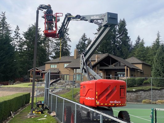 Tennis Court Lighting - LED Upgrade and Retrofit - Landscape Preservation - We installed two new light poles and retrofitted modern LED fixtures onto two existing poles to provide a fully illuminated tennis court for a Lake Tapps waterfront estate. The project required a complete redesign of the lighting layout to improve performance while eliminating the light pollution that had previously reflected across the lake and into neighboring properties.
Scope of Work
Our work included the full design and layout of the electrical circuitry, conduit routing, and lighting configuration. The scope covered:
• Interception and rerouting of existing underground electrical circuits
• Excavation for new underground conduit
• Drilling for light pole bases
• Construction of concrete forms and rebar cages for pole footings
• Concrete placement for all new footings
• Light pole erection and alignment
• Wire installation, pole arm installation, and final fixture installation
Unique Project & Jobsite Challenges
The customer requested two additional tennis court lights within a fully landscaped, mature waterfront backyard with extremely limited equipment access. Existing fixtures had been installed in a way that caused significant glare across Lake Tapps, affecting neighbors and boaters. Our task was to deliver high performance lighting while eliminating unwanted spill light.
We utilized LED fixtures with integral reflectors engineered to direct light downward and slightly forward, ensuring a bright, even playing surface with no glare beyond the court boundaries. This design provides a more pleasant experience for guests, neighbors, and anyone on the water.
To protect the property’s mature landscaping, we used ultra compact excavation equipment for trenching and drilling. Plywood ramps and pathways were installed to safeguard curbing, turf, and vegetation. Because the court was inaccessible to concrete trucks or pump booms, additional crew members transported concrete by wheelbarrow to each footing location. Plywood pathways were also placed over the tennis court surface to allow safe boom lift access for pole installation.
Despite the tight access and sensitive environment, all earthwork, concrete placement, pole installation, and fixture retrofits were completed without damage to the customer’s landscaping, aside from the minimal disturbance required for underground conduit.
Result
The final product is a beautifully illuminated tennis court with precise, low glare lighting and minimal impact to the surrounding property. The upgraded system enhances nighttime play while respecting the privacy and comfort of neighbors, a result that exceeded the customer’s expectations. Tennis Court Lighting - LED Upgrade and Retrofit - Landscape Preservation - We installed two new light poles and retrofitted modern LED fixtures onto two existing poles to provide a fully illuminated tennis court for a Lake Tapps waterfront estate. The project required a complete redesign of the lighting layout to improve performance while eliminating the light pollution that had previously reflected across the lake and into neighboring properties.
Scope of Work
Our work included the full design and layout of the electrical circuitry, conduit routing, and lighting configuration. The scope covered:
• Interception and rerouting of existing underground electrical circuits
• Excavation for new underground conduit
• Drilling for light pole bases
• Construction of concrete forms and rebar cages for pole footings
• Concrete placement for all new footings
• Light pole erection and alignment
• Wire installation, pole arm installation, and final fixture installation
Unique Project & Jobsite Challenges
The customer requested two additional tennis court lights within a fully landscaped, mature waterfront backyard with extremely limited equipment access. Existing fixtures had been installed in a way that caused significant glare across Lake Tapps, affecting neighbors and boaters. Our task was to deliver high performance lighting while eliminating unwanted spill light.
We utilized LED fixtures with integral reflectors engineered to direct light downward and slightly forward, ensuring a bright, even playing surface with no glare beyond the court boundaries. This design provides a more pleasant experience for guests, neighbors, and anyone on the water.
To protect the property’s mature landscaping, we used ultra compact excavation equipment for trenching and drilling. Plywood ramps and pathways were installed to safeguard curbing, turf, and vegetation. Because the court was inaccessible to concrete trucks or pump booms, additional crew members transported concrete by wheelbarrow to each footing location. Plywood pathways were also placed over the tennis court surface to allow safe boom lift access for pole installation.
Despite the tight access and sensitive environment, all earthwork, concrete placement, pole installation, and fixture retrofits were completed without damage to the customer’s landscaping, aside from the minimal disturbance required for underground conduit.
Result
The final product is a beautifully illuminated tennis court with precise, low glare lighting and minimal impact to the surrounding property. The upgraded system enhances nighttime play while respecting the privacy and comfort of neighbors, a result that exceeded the customer’s expectations.