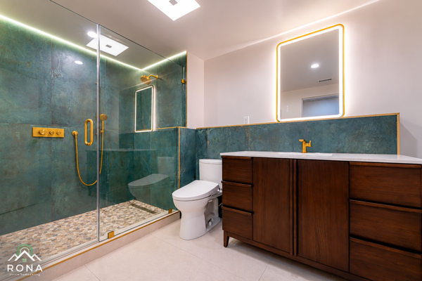 Rosemead Luxurious Teal and Brass Bathroom - Experience the perfect blend of bold color and modern luxury with this recent bathroom remodel by Rona Home Remodeling. This transformation turns an everyday bathroom into a sophisticated, high-end retreat.
Project Highlights:
Striking Custom Tilework: The design is anchored by gorgeous, textured teal wall tiles that run through the shower and act as a bold vanity backsplash, evoking the rich, organic look of oxidized copper or malachite.
Elegant Brass Accents: Premium brushed brass fixtures—including the rain showerhead, hand wand, temperature controls, vanity faucet, and mirror frame—provide a warm, opulent contrast to the deep teal backdrop.
Spa-Inspired Walk-In Shower: The spacious shower area features a sleek, frameless glass enclosure, a natural stone pebble mosaic floor, and integrated LED lighting to create a true spa-like atmosphere.
Modern Vanity & Lighting: A rich, dark wood vanity offers ample storage and warmth, beautifully complemented by a crisp white countertop and an illuminated, smart LED mirror.
Clean, Bright Finishes: A modern, low-profile white toilet and light, large-format floor tiles balance the bold colors, keeping the room feeling expansive and bright. Rosemead Luxurious Teal and Brass Bathroom - Experience the perfect blend of bold color and modern luxury with this recent bathroom remodel by Rona Home Remodeling. This transformation turns an everyday bathroom into a sophisticated, high-end retreat.
Project Highlights:
Striking Custom Tilework: The design is anchored by gorgeous, textured teal wall tiles that run through the shower and act as a bold vanity backsplash, evoking the rich, organic look of oxidized copper or malachite.
Elegant Brass Accents: Premium brushed brass fixtures—including the rain showerhead, hand wand, temperature controls, vanity faucet, and mirror frame—provide a warm, opulent contrast to the deep teal backdrop.
Spa-Inspired Walk-In Shower: The spacious shower area features a sleek, frameless glass enclosure, a natural stone pebble mosaic floor, and integrated LED lighting to create a true spa-like atmosphere.
Modern Vanity & Lighting: A rich, dark wood vanity offers ample storage and warmth, beautifully complemented by a crisp white countertop and an illuminated, smart LED mirror.
Clean, Bright Finishes: A modern, low-profile white toilet and light, large-format floor tiles balance the bold colors, keeping the room feeling expansive and bright.