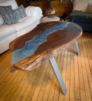 Live Edge Red Walnut River Table - The client requested a captivating red walnut river table that showcases a unique fusion of natural wood and industrial design. Central to their vision was the preservation of the original organic appearance of the red walnut slab, allowing its rich, warm tones and intricate grain patterns to take center stage.
To achieve this, we crafted a sturdy, aesthetically pleasing frame and legs from rectangular metal tubing, each measuring 3 inches in width and 1/2 inch in thickness. The metal was meticulously painted in a sleek metallic silver finish, as per the client's specifications, providing a striking contrast to the warm hues of the walnut.
To enhance the rustic yet modern flair, we incorporated swirls of blue and white epoxy fill, delicately flowing alongside the natural contours of the wood, reminiscent of a serene river cutting through a landscape.
The table was completed with a high-quality wood finishing wax, which not only enriched the color and depth of the walnut but also ensured a silky-smooth surface. After careful buffing, the final result was a stunningly elegant table that embodies both sophistication and natural beauty, destined to be a captivating centerpiece in any space. Live Edge Red Walnut River Table - The client requested a captivating red walnut river table that showcases a unique fusion of natural wood and industrial design. Central to their vision was the preservation of the original organic appearance of the red walnut slab, allowing its rich, warm tones and intricate grain patterns to take center stage.
To achieve this, we crafted a sturdy, aesthetically pleasing frame and legs from rectangular metal tubing, each measuring 3 inches in width and 1/2 inch in thickness. The metal was meticulously painted in a sleek metallic silver finish, as per the client's specifications, providing a striking contrast to the warm hues of the walnut.
To enhance the rustic yet modern flair, we incorporated swirls of blue and white epoxy fill, delicately flowing alongside the natural contours of the wood, reminiscent of a serene river cutting through a landscape.
The table was completed with a high-quality wood finishing wax, which not only enriched the color and depth of the walnut but also ensured a silky-smooth surface. After careful buffing, the final result was a stunningly elegant table that embodies both sophistication and natural beauty, destined to be a captivating centerpiece in any space.