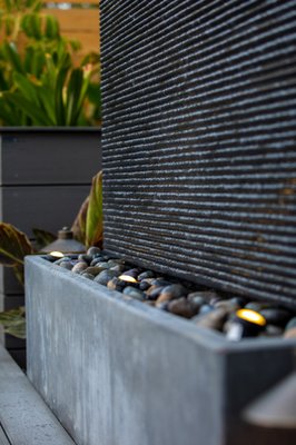 Small Space Social Backyard Design - This narrow property was designed to include a charming social area complete with a fire pit, chairs, and backdrop water feature. In addition, drought-tolerant landscaping surrounding the design decreases noise and adds lush, oasis-like seclusion. The front of the house included a multi-material driveway to draw attention to the best aspects of the house.