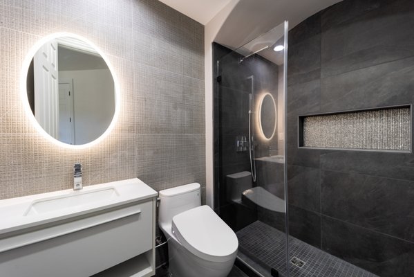 Modern Bathroom with Dark Accents - This modern bathroom renovation features a striking contrast of light and dark elements. The dark gray tile creates a sophisticated and moody atmosphere, while the illuminated round mirror adds a touch of elegance. The frameless glass shower door and sleek fixtures complete the modern aesthetic.