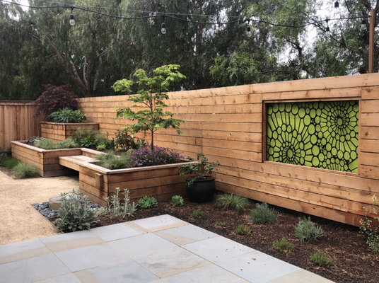 Contemporary Courtyard Garden - The client wanted to transform the space into a contemporary, inviting garden while creating greater privacy from the neighboring school. A new horizontal wood fence was installed to screen the property and provide a warm, contemporary backdrop for the garden. Custom raised planting beds were designed at varying heights to add structure and dimension, with a built-in bench placed between them to create a comfortable spot to sit and enjoy the space.
Connecticut bluestone was selected for the patio, bringing a clean, refined finish that complements the natural warmth of the wood. To make the fence more visually interesting, I designed a framed cutout featuring a custom laser-cut decorative panel. The panel adds color, texture, and a unique artistic focal point while maintaining the privacy the client needed. Layered planting, ornamental grasses, and a Japanese maple soften the modern lines and create a balanced garden that feels both functional and welcoming. Contemporary Courtyard Garden - The client wanted to transform the space into a contemporary, inviting garden while creating greater privacy from the neighboring school. A new horizontal wood fence was installed to screen the property and provide a warm, contemporary backdrop for the garden. Custom raised planting beds were designed at varying heights to add structure and dimension, with a built-in bench placed between them to create a comfortable spot to sit and enjoy the space.
Connecticut bluestone was selected for the patio, bringing a clean, refined finish that complements the natural warmth of the wood. To make the fence more visually interesting, I designed a framed cutout featuring a custom laser-cut decorative panel. The panel adds color, texture, and a unique artistic focal point while maintaining the privacy the client needed. Layered planting, ornamental grasses, and a Japanese maple soften the modern lines and create a balanced garden that feels both functional and welcoming.