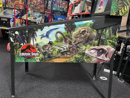 JURASSIC PARK Pinball Machine - Jurassic Park comes with a raptor pen and target, molded t-rex breaking through electric fence, left t-rex ramp, left raptor tower half pipe steel and wire ramp, 3 up post (left return lane, left control room, orbit to divert into pop bumpers) This playfield also has a static helicopter blade, spinning kenetic newton ball jungle explorer vehicle, right spiral steel and wire pteranodon ramp leading back to upper flipper and playfield padlock map that inserts for players to traverse, culminating in 3 mini jackpots and a super jackpot. 

​

A premium adds an interactive, ball throwing dinosaur head. 6 balls installed.

​