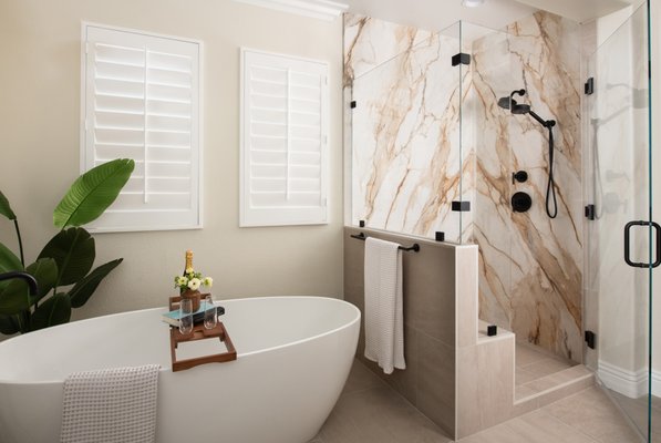 Timeless Mission Viejo Primary Bath and Laundry Room Remodel - The ‘Master Bathroom’ is a space that has only been around for about 40 years. It has evolved dramatically throughout the decades and has only recently become the place where we try to find ultimate tranquility. From color schemes to materials to functionality, every element must work together to create a harmonious design. 
A Primary Bathroom should be hygienic and functional but also calming and relaxing. This light and clean Primary Bathroom design in Mission Viejo creates an illusion that the small bathroom looks bigger than it is. Generously sized vanity mirrors reflect upon the light cream-colored walls, enhancing the perception of the space. The pearl white Omega cabinetry brightens the room with polished quartz stone countertops in Cygnet Drift.  Bold and trendy matte black accents create a striking contrast while also adding a pop of color.  Transitional with a hint of glam, the vanity lights feature a scroll shaped body mounted vertically on both sides of the mirrors.
The star of the bathroom is the book-matched slabs of porcelain on the shower wall, showing proof of amazing craftsmanship. Lining up the veining not only enhances the space but also adds visual interest. Using a glass enclosure allows for an unobstructed view as well as an open and airy feel. A shower niche and bench were also included in a porcelain tile called Morningside, that trickles down to the shower flooring and throughout the bathroom. Enhancing the soothing mood is a freestanding soaking tub featuring a luxurious in-floor tub filler. 
Keeping it simple, a monochromatic color palette was used to create a soothing yet stylish laundry room. To create more storage space, pearl white Omega shaker cabinets with brushed nickel knobs were added above the washer and dryer.  The undermount sink is set into a marble-like polished quartz countertop on an Omega vanity featuring a matte black, single handled pull-down prep faucet. Low maintenance and easy to clean, bacteria resistant glazed porcelain tiles add a modern touch in a hexagon shape. 
If you’re planning your own Primary Bathroom remodel in Mission Viejo or any other home remodeling project in Orange County, Sea Pointe Design & Remodel has you covered! We’ve been meeting the remodeling needs of Orange County residents for over 35 years and we’d love to remodel YOUR home next. Simply give us a call at (949) 749-5642 or fill out the form below to schedule your complimentary design consultation.