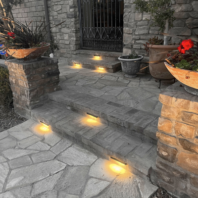 Landscape Lighting - Hardscaping plays a big role in how attractive and live-able an outdoor space is. But so does its lighting - those lovely stairs and inviting patio of yours will be worthless if consumed by nighttime darkness. Luckily, there's hardscape lighting. This article will introduce you to it, go over its many types, and explain how you can incorporate it into your outdoor space.
What Is Hardscape Lighting?
As implied by the name, hardscape lighting is a type of exterior lighting specifically designed to be installed in and used within hardscapes—any man-made structures found in outdoor spaces, such as patios, walkways and retaining walls.
Hardscape Lighting is a popular way to create drama in outdoor spaces and increase security by illuminating entrances and other boundaries. It’s also an ideal solution for areas without vegetation, like driveways and garages. The light will bounce off the walls and other hard surfaces, enabling a softer but more even illumination that can’t be achieved with conventional bulbs or lanterns.
Hardscape Lighting is available in a variety of colors, sizes, and shapes to match any hardscape project. It can be installed as standalone fixtures or integrated into larger systems with multiple lights for more dramatic effects.
How Is Hardscape Lighting Different From Traditional Landscape Lighting?
Hardscape lighting, landscape lighting... potato potahto, right?
Not quite. While both are designed to illuminate and enhance outdoor spaces, hardscape lighting has a distinct purpose in illuminating hard surfaces, such as walkways, driveways, patios and other outdoor features. Fixtures are often set into the ground or built into structures like walls, making them permanent, less visible to the eye, and sometimes more complicated to install.
While traditional forms of landscape lighting can also be hard to set up, many products are standalone and require no more than a steak in the ground. In every case with hardscape lighting, you're drilling into concrete or masonry, and possibly wiring the fixtures into dedicated circuits.
Types of Hardscape Lighting
We've already established the one thing all hardscape lights have in common - hard surface illumination. Beyond that, products can be split up into several categories, and then further into various shapes, sizes, and styles. Read below for an overview of the biggest types to know.
Wall Lights
Wall lights are the most common type of hardscape lighting. They typically feature flat, rectangular frames mounted onto a wall or other vertical surface that will give off soft light in an area. These can be used on sides of buildings, staircases, and other areas that you want to highlight.
Path Lights
Path lights are used to illuminate a planar surface like a driveways or walkway. The first and foremost reason for their use is safety; path lights provide important visibility to property goers and visitors. On a more decorative note, path lights can also be used to draw more attention to walkways, paths, or their surrounding features.
Spot Lights
In a literal sense, spotlights are the most powerful type of hardscape lighting. They are designed to focus brightness in a single direction and usually throw a far distance. This creates a dramatic beam that can be angled differently for different effects. Spot lights are great for highlighting statues, trees, and other features of a property.
Post Lights
Post lights are most commonly used to provide additional lighting in areas that may be more difficult to reach with wall lights or pathlights (like stairs). They feature a tall upright light post attached by one or two arms on either side and give off soft illumination in an area- perfect for decks, gazebos, and outdoor patios.
Deck Lights
Deck lights are intended for decks but can also be used on steps or other outdoor surfaces like patios and porches. These lights are installed directly into the wood or stone of a surface to give off extra illumination right where you need it.
Step Lights
Step lights are installed directly within the individual steps of an outdoor staircase. Some come in strip form, while others fall into the category of well lights and require a hole to be cut into the step. Either way, they are an effective and attractive way to light a staircase.
Underwater Lights
Underwater LightsUnderwater lighting is great for those who want to feature the beauty of their pool, pond, or other water feature at night. Underwater products are built to withstand long-term submersion and produce an output bright enough to illuminate large and deep spaces.
How Hardscape Lights Can Make Your Yard (and Life) Better
Hardscape lighting brings a different level of sophistication to an outdoor space. Sure, some lanterns here and there can give your property a nice feel, but they won't get the job done like these products can. Functional, beautiful, and practical, properly-installed hardscape lights. Landscape Lighting - Hardscaping plays a big role in how attractive and live-able an outdoor space is. But so does its lighting - those lovely stairs and inviting patio of yours will be worthless if consumed by nighttime darkness. Luckily, there's hardscape lighting. This article will introduce you to it, go over its many types, and explain how you can incorporate it into your outdoor space.
What Is Hardscape Lighting?
As implied by the name, hardscape lighting is a type of exterior lighting specifically designed to be installed in and used within hardscapes—any man-made structures found in outdoor spaces, such as patios, walkways and retaining walls.
Hardscape Lighting is a popular way to create drama in outdoor spaces and increase security by illuminating entrances and other boundaries. It’s also an ideal solution for areas without vegetation, like driveways and garages. The light will bounce off the walls and other hard surfaces, enabling a softer but more even illumination that can’t be achieved with conventional bulbs or lanterns.
Hardscape Lighting is available in a variety of colors, sizes, and shapes to match any hardscape project. It can be installed as standalone fixtures or integrated into larger systems with multiple lights for more dramatic effects.
How Is Hardscape Lighting Different From Traditional Landscape Lighting?
Hardscape lighting, landscape lighting... potato potahto, right?
Not quite. While both are designed to illuminate and enhance outdoor spaces, hardscape lighting has a distinct purpose in illuminating hard surfaces, such as walkways, driveways, patios and other outdoor features. Fixtures are often set into the ground or built into structures like walls, making them permanent, less visible to the eye, and sometimes more complicated to install.
While traditional forms of landscape lighting can also be hard to set up, many products are standalone and require no more than a steak in the ground. In every case with hardscape lighting, you're drilling into concrete or masonry, and possibly wiring the fixtures into dedicated circuits.
Types of Hardscape Lighting
We've already established the one thing all hardscape lights have in common - hard surface illumination. Beyond that, products can be split up into several categories, and then further into various shapes, sizes, and styles. Read below for an overview of the biggest types to know.
Wall Lights
Wall lights are the most common type of hardscape lighting. They typically feature flat, rectangular frames mounted onto a wall or other vertical surface that will give off soft light in an area. These can be used on sides of buildings, staircases, and other areas that you want to highlight.
Path Lights
Path lights are used to illuminate a planar surface like a driveways or walkway. The first and foremost reason for their use is safety; path lights provide important visibility to property goers and visitors. On a more decorative note, path lights can also be used to draw more attention to walkways, paths, or their surrounding features.
Spot Lights
In a literal sense, spotlights are the most powerful type of hardscape lighting. They are designed to focus brightness in a single direction and usually throw a far distance. This creates a dramatic beam that can be angled differently for different effects. Spot lights are great for highlighting statues, trees, and other features of a property.
Post Lights
Post lights are most commonly used to provide additional lighting in areas that may be more difficult to reach with wall lights or pathlights (like stairs). They feature a tall upright light post attached by one or two arms on either side and give off soft illumination in an area- perfect for decks, gazebos, and outdoor patios.
Deck Lights
Deck lights are intended for decks but can also be used on steps or other outdoor surfaces like patios and porches. These lights are installed directly into the wood or stone of a surface to give off extra illumination right where you need it.
Step Lights
Step lights are installed directly within the individual steps of an outdoor staircase. Some come in strip form, while others fall into the category of well lights and require a hole to be cut into the step. Either way, they are an effective and attractive way to light a staircase.
Underwater Lights
Underwater LightsUnderwater lighting is great for those who want to feature the beauty of their pool, pond, or other water feature at night. Underwater products are built to withstand long-term submersion and produce an output bright enough to illuminate large and deep spaces.
How Hardscape Lights Can Make Your Yard (and Life) Better
Hardscape lighting brings a different level of sophistication to an outdoor space. Sure, some lanterns here and there can give your property a nice feel, but they won't get the job done like these products can. Functional, beautiful, and practical, properly-installed hardscape lights.