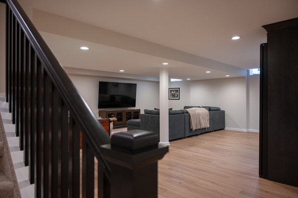 Ann Arbor Finished Basement - This finished basement in Ann Arbor, Michigan combines comfort, function, and thoughtful design throughout. The space features a spacious living room, partial basement bar with wine coolers, a finished bathroom, wood flooring, a drywall ceiling, and a dedicated area for working out. Even the practical details were designed to blend in, including a hidden sump pump cover that keeps essential equipment accessible without taking away from the finished look. Completed by the Basements Plus team, this basement gives the homeowners more space to relax, entertain, and enjoy their home every day.