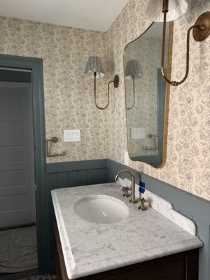 Elegant Powder Room Remodel in Ocean City, NJ - Matera Builders transformed this powder room in Ocean City, NJ into a warm, classic space with the character of a historic home and the durability a busy shore house needs. The goal was a small room with big personality: rich color, layered pattern, and fixtures that feel collected rather than ordered from a catalog.

Design and finishes

The walls pair a soft floral wallpaper in blue-gray on cream with painted V-groove wainscoting below. The wainscoting is finished in a deep blue-green and capped with a matching chair rail, which carries the color around the room and protects the lower walls from everyday wear. Window and door trim are painted to match, giving the room a finished, custom look.

The centerpiece is a furniture-style vanity with turned legs, an open lower shelf and a dark wood finish. It's topped with a white marble countertop with a shaped backsplash and an oval undermount sink. A brass widespread faucet with cross handles sits alongside brass sconces with pleated patterned shades, a curved brass-framed mirror, and a matching brass towel bar and toilet paper holder.

Underfoot, a patterned tile floor in warm terracotta and white adds a bold geometric contrast to the floral walls. A comfort-height toilet and an exhaust fan finish the room, keeping it practical for guests and family alike.

What this project included

Full powder room remodel
Wallpaper installation
V-groove wainscoting and chair rail installation and painting
Furniture-style vanity and marble top installation
Undermount sink and brass faucet installation
Wall sconce and mirror installation
Patterned tile flooring
Toilet and bath accessory installation
Trim carpentry and finish painting

Why homeowners choose Matera for powder rooms

A powder room is the bathroom your guests see most, which makes it one of the best places to add design impact. Because the space is small, every detail shows, from how the wallpaper pattern lines up at the corners to how cleanly the wainscoting meets the trim. Our crews handle the carpentry, tile, fixtures and finish work so the room comes together as one design rather than a collection of separate trades.

Frequently asked questions

Who does powder room remodels in Ocean City, NJ?
Matera Builders remodels powder rooms, bathrooms and kitchens for homeowners in Ocean City, Avalon, Stone Harbor and Sea Isle City, NJ. We've served the South Jersey shore since 1988.

Can you install wallpaper and wainscoting as part of a bathroom remodel?
Yes. Wallpaper, wainscoting, chair rail and trim carpentry are part of our remodeling work, so they're coordinated with the vanity, tile and fixtures from the start.

When is the best time to remodel a shore home bathroom?
The fall and winter off-season is ideal. Homes are often empty, work doesn't interrupt summer use or rentals, and the project is finished before the season starts.

Does Matera Builders only install windows and doors?
No. Matera Builders is a full home remodeling contractor and an Andersen Elite Certified window and door installer. We handle kitchen and bath remodels, flooring, siding, finish carpentry and more.