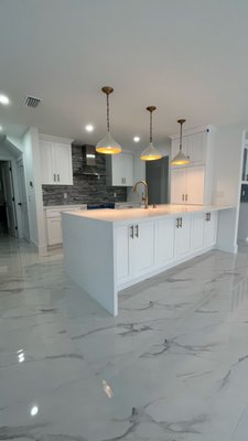 Modern White Kitchen Remodel - This kitchen remodel embodies elegance, light, and functionality. Featuring sleek white cabinetry, marble-look flooring, and brass accents, the space achieves a perfect balance between warmth and modern sophistication. The open layout enhances natural light, while the statement island and custom backsplash add depth and character. Every finish was carefully selected to create a timeless, inviting kitchen designed for everyday living and unforgettable gatherings.