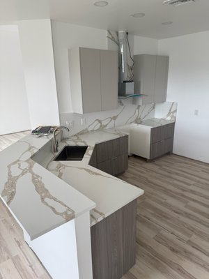 Modern Wood Tone Warm Color Kitchen - This stunning modern kitchen showcases a sophisticated two-tone design, pairing light taupe upper cabinets with warm textured wood lower cabinetry for a perfect balance of sleek and natural elements. Striking waterfall-edge countertops and full-height backsplash in a bold white quartz with dramatic veining create a seamless, luxurious flow across the space. The clean lines, minimalist hardware, and integrated vent hood emphasize the contemporary aesthetic, while the light wood-look flooring ties the entire look together with a soft, welcoming feel. Thoughtfully designed for both style and functionality, this kitchen offers an elegant yet inviting space perfect for modern living. Modern Wood Tone Warm Color Kitchen - This stunning modern kitchen showcases a sophisticated two-tone design, pairing light taupe upper cabinets with warm textured wood lower cabinetry for a perfect balance of sleek and natural elements. Striking waterfall-edge countertops and full-height backsplash in a bold white quartz with dramatic veining create a seamless, luxurious flow across the space. The clean lines, minimalist hardware, and integrated vent hood emphasize the contemporary aesthetic, while the light wood-look flooring ties the entire look together with a soft, welcoming feel. Thoughtfully designed for both style and functionality, this kitchen offers an elegant yet inviting space perfect for modern living.