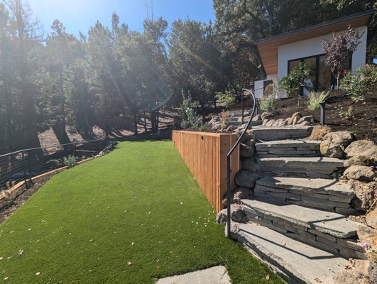 Landscaped Hillside with Studio - Steep, unusable hillside transformed with terraced retaining walls to create two large usable spaces - an artificial turf area and a studio. Features such as Redwood horizontal slats, flagstone cap, and rock help to add interest to the walls. Drought tolerant planting will eventually grow in and further soften the profile. Stainless steel cable railing keeps it minimal and clean. The studio features tall windows and bifold doors, to maximize light and views. Efficient low voltage LED lighting highlights the landscape features in the evening. Landscaped Hillside with Studio - Steep, unusable hillside transformed with terraced retaining walls to create two large usable spaces - an artificial turf area and a studio. Features such as Redwood horizontal slats, flagstone cap, and rock help to add interest to the walls. Drought tolerant planting will eventually grow in and further soften the profile. Stainless steel cable railing keeps it minimal and clean. The studio features tall windows and bifold doors, to maximize light and views. Efficient low voltage LED lighting highlights the landscape features in the evening.