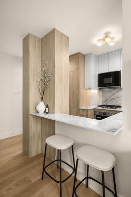 Full Home Remodeling NYC - This bright and modern renovation delivers a refined, Scandinavian-inspired aesthetic. In the kitchen, a warm wood base blends beautifully with cool gray upper cabinets for a fresh and calming palette. Sleek slab-front cabinetry, minimalist hardware, and continuous-vein quartz surfaces on the counters and backsplash give the space a polished and seamless feel. Under-cabinet lighting and integrated appliances ensure both form and function, while the passthrough breakfast bar enhances the apartment’s flow and sociability.
The bathroom continues the same sophisticated tone with large-format marble-look porcelain tiles paired with soft beige stone accents. A walk-in glass shower features recessed shelving, a rain showerhead, and a linear drain for a modern, barrier-free design. The wall-mounted vanity, contemporary fixtures, and mirrored medicine cabinet with integrated lighting round out the bathroom's serene, spa-like feel—balancing simplicity, warmth, and high-end detail throughout. Full Home Remodeling NYC - This bright and modern renovation delivers a refined, Scandinavian-inspired aesthetic. In the kitchen, a warm wood base blends beautifully with cool gray upper cabinets for a fresh and calming palette. Sleek slab-front cabinetry, minimalist hardware, and continuous-vein quartz surfaces on the counters and backsplash give the space a polished and seamless feel. Under-cabinet lighting and integrated appliances ensure both form and function, while the passthrough breakfast bar enhances the apartment’s flow and sociability.
The bathroom continues the same sophisticated tone with large-format marble-look porcelain tiles paired with soft beige stone accents. A walk-in glass shower features recessed shelving, a rain showerhead, and a linear drain for a modern, barrier-free design. The wall-mounted vanity, contemporary fixtures, and mirrored medicine cabinet with integrated lighting round out the bathroom's serene, spa-like feel—balancing simplicity, warmth, and high-end detail throughout.