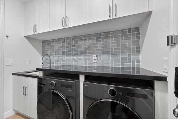 Style Laundry Retreat with Custom Storage - This beautifully designed laundry room combines style and functionality with sleek custom cabinetry, modern black appliances, and a sophisticated gray tile backsplash. Thoughtfully designed storage solutions maximize every inch of space, while the durable countertops and elegant flooring create a clean, contemporary feel. Bright lighting and a neutral color palette make this laundry area both practical and inviting—perfect for everyday organization and efficiency.
