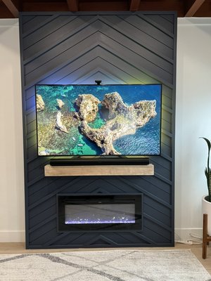 Fireplace project - Before: The living room featured a plain, white wall with minimal decoration. The space lacked a focal point and had a somewhat cluttered and unfinished appearance. Despite the presence of some plants and household items, the overall aesthetic was unremarkable, and the room felt in need of a stylish upgrade.
After: The transformation is striking, with the once plain wall now boasting a stunning custom accent wall. The new design includes a sleek, dark geometric pattern that adds depth and elegance to the room. Integrated into the accent wall are a mounted TV and a modern electric fireplace, creating a sophisticated and functional focal point. The addition of ambient lighting enhances the overall atmosphere, making the living room a visually appealing and cozy environment. Fireplace project - Before: The living room featured a plain, white wall with minimal decoration. The space lacked a focal point and had a somewhat cluttered and unfinished appearance. Despite the presence of some plants and household items, the overall aesthetic was unremarkable, and the room felt in need of a stylish upgrade.
After: The transformation is striking, with the once plain wall now boasting a stunning custom accent wall. The new design includes a sleek, dark geometric pattern that adds depth and elegance to the room. Integrated into the accent wall are a mounted TV and a modern electric fireplace, creating a sophisticated and functional focal point. The addition of ambient lighting enhances the overall atmosphere, making the living room a visually appealing and cozy environment.