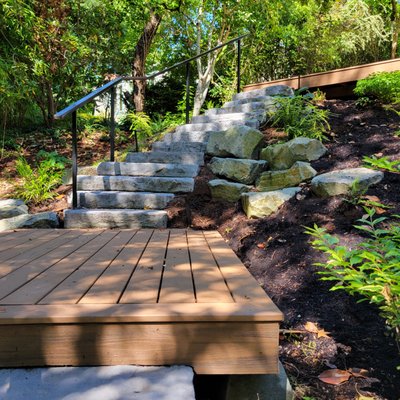Scenic Descent: A Safe and Stunning Lakeside Path - The landscape design creates a safe and inviting transition from the home to the lakeside patio. Castohn irregular steps wind gracefully down the slope, their natural stone-like appearance complementing the surrounding environment. Trex decking platforms provide comfortable resting points along the way, allowing for moments of pause to take in the scenic views. A custom metal railing adds both safety and elegance, its sleek design subtly enhancing the natural surroundings without obstructing the view.
Carefully selected plantings create a lush, naturalistic setting, ensuring year-round interest while stabilizing the hillside. A mix of shrubs, trees, and perennials softens the edges of the stairs and viewing decks, offering seasonal color, texture, and movement. As they mature, these plantings will weave together into a rich tapestry, providing habitat for wildlife and preventing erosion on the sloped terrain.
The addition of a dry stream bed, starting at a natural spring source and winding down to the lake's edge, helps ensure water containment while providing another eye-catching feature. This naturalistic element enhances the landscape’s visual appeal and aids in directing runoff efficiently, preventing erosion and integrating seamlessly with the plantings.
The result is a thoughtfully designed landscape that harmonizes safety, aesthetics, and ecological function, inviting visitors to meander down the path and fully enjoy the lakeside retreat. Scenic Descent: A Safe and Stunning Lakeside Path - The landscape design creates a safe and inviting transition from the home to the lakeside patio. Castohn irregular steps wind gracefully down the slope, their natural stone-like appearance complementing the surrounding environment. Trex decking platforms provide comfortable resting points along the way, allowing for moments of pause to take in the scenic views. A custom metal railing adds both safety and elegance, its sleek design subtly enhancing the natural surroundings without obstructing the view.
Carefully selected plantings create a lush, naturalistic setting, ensuring year-round interest while stabilizing the hillside. A mix of shrubs, trees, and perennials softens the edges of the stairs and viewing decks, offering seasonal color, texture, and movement. As they mature, these plantings will weave together into a rich tapestry, providing habitat for wildlife and preventing erosion on the sloped terrain.
The addition of a dry stream bed, starting at a natural spring source and winding down to the lake's edge, helps ensure water containment while providing another eye-catching feature. This naturalistic element enhances the landscape’s visual appeal and aids in directing runoff efficiently, preventing erosion and integrating seamlessly with the plantings.
The result is a thoughtfully designed landscape that harmonizes safety, aesthetics, and ecological function, inviting visitors to meander down the path and fully enjoy the lakeside retreat.