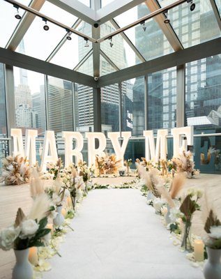 Rooftop Proposal Packages - Elevate your love story to new heights with Enchanting Engagements in the heart of New York City. Our rooftop proposal packages, meticulously curated by our expert team, stand out as some of our most beloved offerings. Immerse yourself in the unparalleled beauty of the cityscape, a view that's truly one-of-a-kind.
Our thoughtfully crafted package eliminates the need for you to fret over the details; we've taken care of it all. Still, we leave room for your personal touch and customization, ensuring that your proposal is a reflection of your unique love story.
The stars of the show are our striking two and three-foot letters, stealing the spotlight with their elegance and charm. But we go the extra mile by infusing your proposal with personal touches. Pictures and songs that hold a special place in your hearts are seamlessly woven into the experience, making it an unforgettable chapter in your love story.
Whether you dream of an indoor or outdoor setting, we've got you covered with a range of options tailored to your budget. Our mission is to make your vision come to life, and our team specializes in crafting unique proposal ideas and providing impeccable planning services right here in the heart of New York City.
Picture you and your partner basking in the romance of a rooftop in the Big Apple, adorned with exquisite decorations and blooming with fresh flowers. At Enchanting Engagements, we're here to turn this dream into a reality. Our rooftop proposal packages are highly customizable, allowing us to create a moment that you and your loved one will treasure forever. Let us be the architects of your unforgettable proposal in the city that never sleeps. Rooftop Proposal Packages - Elevate your love story to new heights with Enchanting Engagements in the heart of New York City. Our rooftop proposal packages, meticulously curated by our expert team, stand out as some of our most beloved offerings. Immerse yourself in the unparalleled beauty of the cityscape, a view that's truly one-of-a-kind.
Our thoughtfully crafted package eliminates the need for you to fret over the details; we've taken care of it all. Still, we leave room for your personal touch and customization, ensuring that your proposal is a reflection of your unique love story.
The stars of the show are our striking two and three-foot letters, stealing the spotlight with their elegance and charm. But we go the extra mile by infusing your proposal with personal touches. Pictures and songs that hold a special place in your hearts are seamlessly woven into the experience, making it an unforgettable chapter in your love story.
Whether you dream of an indoor or outdoor setting, we've got you covered with a range of options tailored to your budget. Our mission is to make your vision come to life, and our team specializes in crafting unique proposal ideas and providing impeccable planning services right here in the heart of New York City.
Picture you and your partner basking in the romance of a rooftop in the Big Apple, adorned with exquisite decorations and blooming with fresh flowers. At Enchanting Engagements, we're here to turn this dream into a reality. Our rooftop proposal packages are highly customizable, allowing us to create a moment that you and your loved one will treasure forever. Let us be the architects of your unforgettable proposal in the city that never sleeps.