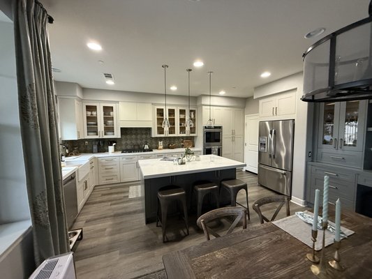 Kitchen Remodel - Rancho Mission Viejo - This remodel transforms the space from a traditional white kitchen into a cleaner, sleeker shaker-style design with a fresh, modern feel.
The original white cabinetry and built‑in desk area have been updated to White shaker doors and drawers with simple, linear hardware. A key feature of the new design is the introduction of greenish-toned cabinets, used on the built‑in hutch/desk area and the large island. This muted green-gray color adds depth and character while still feeling neutral and sophisticated.
The perimeter cabinetry remains light, which keeps the room bright and open, but the contrast of the green cabinetry grounds the space and visually connects the kitchen with the adjacent dining area. Glass-front upper cabinets and open shelving provide display space and help break up the runs of doors, while the streamlined fixtures and finishes—pendant lighting, stainless appliances, and updated backsplash—reinforce the contemporary shaker look.
Overall, the remodel creates a cohesive, updated kitchen with a soft, modern palette where the greenish cabinets become the subtle statement element! Kitchen Remodel - Rancho Mission Viejo - This remodel transforms the space from a traditional white kitchen into a cleaner, sleeker shaker-style design with a fresh, modern feel.
The original white cabinetry and built‑in desk area have been updated to White shaker doors and drawers with simple, linear hardware. A key feature of the new design is the introduction of greenish-toned cabinets, used on the built‑in hutch/desk area and the large island. This muted green-gray color adds depth and character while still feeling neutral and sophisticated.
The perimeter cabinetry remains light, which keeps the room bright and open, but the contrast of the green cabinetry grounds the space and visually connects the kitchen with the adjacent dining area. Glass-front upper cabinets and open shelving provide display space and help break up the runs of doors, while the streamlined fixtures and finishes—pendant lighting, stainless appliances, and updated backsplash—reinforce the contemporary shaker look.
Overall, the remodel creates a cohesive, updated kitchen with a soft, modern palette where the greenish cabinets become the subtle statement element!