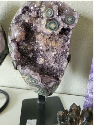 Uruguay Amethyst on a stand - Bring the mystical energy of Uruguay Amethyst into your home with this stunning piece on a stand! This beautiful purple, orange and green geode cluster crystal with several stalactite formations on it, is not only visually captivating, but it is also believed to possess powerful healing properties. Place it in your meditation space or living room to promote peace, balance, and spiritual growth. Perfect for any crystal enthusiast or anyone looking to add a touch of magic to their space. Get your hands on this unique piece today! Uruguay Amethyst on a stand - Bring the mystical energy of Uruguay Amethyst into your home with this stunning piece on a stand! This beautiful purple, orange and green geode cluster crystal with several stalactite formations on it, is not only visually captivating, but it is also believed to possess powerful healing properties. Place it in your meditation space or living room to promote peace, balance, and spiritual growth. Perfect for any crystal enthusiast or anyone looking to add a touch of magic to their space. Get your hands on this unique piece today!