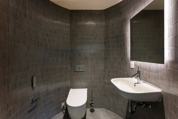 Curved-Wall Bathroom Remodel - This bathroom remodel features a sleek, minimalist design with dark, narrow rectangular tiles covering the walls and floor, creating a unified and modern look. The space includes a wall-mounted toilet and a contemporary, wall-mounted sink with an elegant faucet, both contributing to the streamlined, space-efficient layout. The curved walls add architectural interest, while integrated lighting around the mirror provides a soft, ambient glow. Exposed plumbing beneath the sink adds an industrial touch, and the discreet flush system and hidden electrical outlets maintain the room's clean, uncluttered aesthetic. Overall, the design emphasizes functionality, modern elegance, and simplicity.