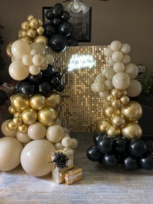 Elegant 47th Birthday Balloon Decor by Sophie’s Balloon Designs - This sophisticated balloon arrangement was created to celebrate a 47th birthday in true style. The design features a luxurious color palette of black, gold, and champagne, exuding elegance and class. The towering balloon garlands, composed of various sizes and metallic finishes, frame a shimmering gold sequin backdrop, complemented by a glowing “Happy Birthday” neon sign for an extra touch of glamour.
The decor is completed with wrapped gold gift boxes, adding a festive and celebratory feel to the setup. This arrangement serves as the perfect centerpiece for photos and creates a memorable atmosphere for an upscale celebration. Elegant 47th Birthday Balloon Decor by Sophie’s Balloon Designs - This sophisticated balloon arrangement was created to celebrate a 47th birthday in true style. The design features a luxurious color palette of black, gold, and champagne, exuding elegance and class. The towering balloon garlands, composed of various sizes and metallic finishes, frame a shimmering gold sequin backdrop, complemented by a glowing “Happy Birthday” neon sign for an extra touch of glamour.
The decor is completed with wrapped gold gift boxes, adding a festive and celebratory feel to the setup. This arrangement serves as the perfect centerpiece for photos and creates a memorable atmosphere for an upscale celebration.