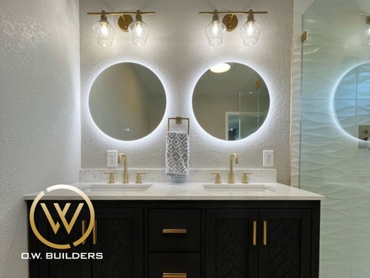 Bathroom Remodel | San Francisco - This stunning remodel combines dark wood cabinetry, brushed brass accents, and wave-textured wall tile for a bold, high-end coastal aesthetic. The dual backlit mirrors, integrated lighting, and frameless shower bring spa-quality comfort and visual symmetry to a space built for both luxury and functionality.