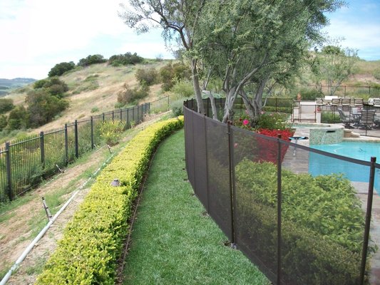 Pool Fence Installed in Grass. Securely installed in grass, providing a strong safety barrier while keeping your yard open and attractive. - In this photo series you will see a complete mesh pool fence installation built directly on a grassy surface. This project highlights the right way to install a fence in grass while keeping the finished look neat and secure.

Site preparation and layout
The process began with careful planning. Before any sleeves or footings were placed, the grass was cut and removed in clean sections to mark where each pole would be installed. This creates a precise line for the fence and prevents damage to the surrounding yard. The layout uses gentle curves instead of sharp angles. Sharp turns are difficult in grass, so smooth curves create a cleaner look and help the installation go more smoothly.

PVC sleeves and concrete footings
PVC sleeves were arranged on the ground to show the exact spots for each fence post. These sleeves guide the placement of the PVC sleeves and concrete work that follows. Once the positions were confirmed, concrete was poured to form solid footings. The sleeves sit inside the concrete, creating a secure foundation for the removable aluminum poles.

Round terminating post in grass
In one section a round terminating post was used to make a 90 degree turn. This type of post makes a clean transition and holds the mesh securely. It also reduces stress on the mesh where the fence changes direction.

In-progress details
Some photos show the job while still in progress. PVC pipe is extending from the freshly poured concrete footings. These pipes mark where the aluminum poles will go and show how the fence structure is anchored. Close up photos show the concrete footings with PVC sleeves set firmly in place. Each sleeve will hold an aluminum pole that supports the mesh.

Fence next to a concrete deck
The photos also show areas where the fence is installed right next to a concrete pool deck. The grass was cut neatly along the edge and the fence lines up flush to the deck. This keeps the fence tight to the pool area and eliminates gaps that could compromise safety.

Mesh and hardware
When the structure is ready, the mesh panels are installed between the poles. All-Safe mesh fencing meets or exceeds ASTM safety standards and is designed to be difficult for children to climb. The spacing between poles is kept under 40 inches and the height meets local code requirements, usually 48 or 60 inches. Self-closing gates, latch hardware, and other safety features are added at the end of the process, with latches placed at least 54 inches above the ground for child safety.

Safety considerations
The bottom of the mesh fence sits close to the ground, less than one inch above the surface. This makes it difficult for children or pets to crawl under the fence. Poles are spaced so that a 4 inch sphere cannot pass between them, which is an important ASTM safety requirement.

Why grass installations need extra care
Installing in grass takes more preparation than installing in concrete alone. The soil must be carefully cut to create stable footing locations and to prevent shifting over time. Gentle curves are preferred over sharp turns because they disturb less grass and help maintain strength.

Progression of the job
- Grass is cut to mark footing spots.
- PVC sleeves are placed to outline the fence.
- Concrete footings are poured around each sleeve.
- Round terminating posts are added to make clean turns.
- Mesh panels are installed between aluminum poles.

Close up views show how sleeves, footings, and hardware come together.

This installation shows how mesh pool fencing can be safely and cleanly installed in grass while maintaining a professional appearance. Each step, from cutting the turf to placing PVC sleeves and pouring concrete, follows the methods recommended by All-Safe. The finished fence provides a strong, removable safety barrier while blending well with the surrounding landscape.
