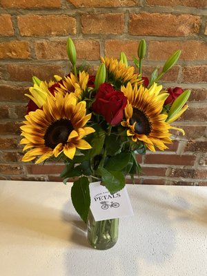 Midnight Love | Summer - Fresh Flower Bouquet with Vase
Chocolate Sunflowers, Midnight Long Stem Roses and Young Yellow Lillies - This cheerful bouquet features bold sunflowers paired with classic red roses and lilies for a mix of warmth and elegance. The one pictured is our Medium size, perfect for a dining table centerpiece or as a gift that makes a strong impression.

We also offer it in:
	•	Small – A charming, compact version ideal for desks, nightstands, or smaller gestures.
	•	Large – A fuller, more dramatic arrangement with additional blooms for maximum impact.
	•	Handbag – Our signature style, arranged in a chic handbag-style base for a modern, fashion-forward presentation.

No matter the size, each bouquet is handcrafted, finished with care, and hand-delivered with a handwritten card—because every detail matters.