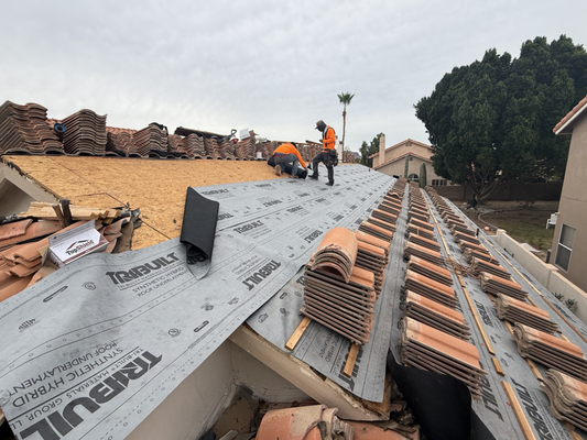 Arizona Tile Roof - Roof replacement in progress featuring a clay tile roofing system. The old tiles have been removed, synthetic underlayment has been installed, and new battens are laid out for proper tile alignment. Stacks of terracotta tiles are staged across the roof, ready for installation, while crew members work on securing the new roofing system.