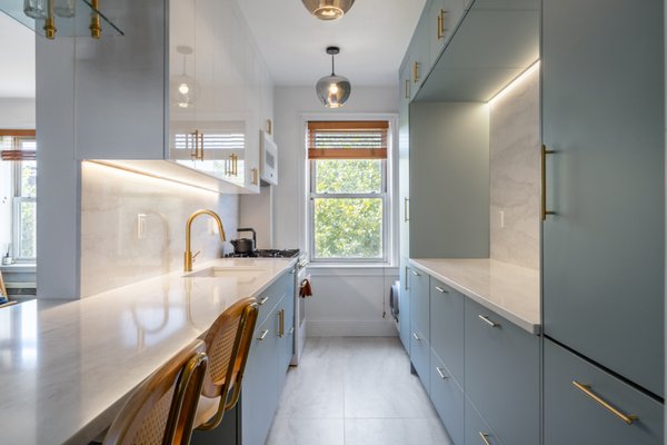 Modern Blue Galley Kitchen Transformation in Queens - This Queens kitchen renovation by NYKB is a refined example of how thoughtful design can completely transform a compact space into something both highly functional and visually elevated.
The layout follows a classic galley configuration, but with a modern, streamlined approach that maximizes every inch. Custom cabinetry in a soft blue tone brings a calm, sophisticated feel, while the slim brass hardware adds just the right amount of warmth and contrast. The cabinetry was extended vertically to increase storage capacity, creating a clean, built-in look that draws the eye upward and enhances the sense of height.
A continuous slab backsplash and countertop in a light, marble-look finish creates a seamless and elegant surface, reflecting natural light from the window and helping the space feel brighter and more open. The integrated under-cabinet lighting adds both function and ambiance, highlighting the material and providing practical task lighting.
On one side, a custom peninsula was introduced to serve as a casual seating area, making the kitchen more interactive and suitable for everyday living. The addition of bar seating with natural wood and woven textures softens the modern lines and brings in an inviting, organic element.
Lighting was carefully selected to complement the overall aesthetic. The glass pendant fixtures add a contemporary touch without overwhelming the space, keeping the design light and airy.
Overall, this project balances modern minimalism with warmth and practicality. It reflects NYKB’s approach of combining smart space planning, high-quality materials, and clean design to create kitchens that are not only beautiful but truly livable. Modern Blue Galley Kitchen Transformation in Queens - This Queens kitchen renovation by NYKB is a refined example of how thoughtful design can completely transform a compact space into something both highly functional and visually elevated.
The layout follows a classic galley configuration, but with a modern, streamlined approach that maximizes every inch. Custom cabinetry in a soft blue tone brings a calm, sophisticated feel, while the slim brass hardware adds just the right amount of warmth and contrast. The cabinetry was extended vertically to increase storage capacity, creating a clean, built-in look that draws the eye upward and enhances the sense of height.
A continuous slab backsplash and countertop in a light, marble-look finish creates a seamless and elegant surface, reflecting natural light from the window and helping the space feel brighter and more open. The integrated under-cabinet lighting adds both function and ambiance, highlighting the material and providing practical task lighting.
On one side, a custom peninsula was introduced to serve as a casual seating area, making the kitchen more interactive and suitable for everyday living. The addition of bar seating with natural wood and woven textures softens the modern lines and brings in an inviting, organic element.
Lighting was carefully selected to complement the overall aesthetic. The glass pendant fixtures add a contemporary touch without overwhelming the space, keeping the design light and airy.
Overall, this project balances modern minimalism with warmth and practicality. It reflects NYKB’s approach of combining smart space planning, high-quality materials, and clean design to create kitchens that are not only beautiful but truly livable.