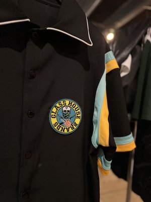Custom Shirts & More - A stylish black button-up shirt featuring the distinctive "Glass House Brands" logos, all constructed at The Embroidery Shop. Accentuated with light blue and yellow stripes on the sleeves, it adds a pop of color. The white piping on the collar enhances its crisp, clean finish. This is just an example of all that we can do at The Embroidery Shop. Visit us to discover how we can create unique apparel for you!