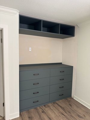 Mineral Green Dresser with Matte Finish - What was an open clothes niche with a single hanging rod, shoe racks and no power planning - a storage-only corner. We converted the space into a built-in TV/media wall: a full-width cabinets with eight deep drawers for concealed storage, open upper cubbies for decor elements, and a center power outlet ready for the TV. A matte deep-green finish and sleek black pulls are turning a rough clothes area into a calm, high-end media zone that organizes the room and elevates the look. Mineral Green Dresser with Matte Finish - What was an open clothes niche with a single hanging rod, shoe racks and no power planning - a storage-only corner. We converted the space into a built-in TV/media wall: a full-width cabinets with eight deep drawers for concealed storage, open upper cubbies for decor elements, and a center power outlet ready for the TV. A matte deep-green finish and sleek black pulls are turning a rough clothes area into a calm, high-end media zone that organizes the room and elevates the look.