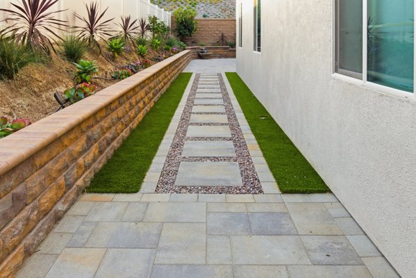 Custom Paver Patio, Retaining Wall & Landscape Lighting Design - This backyard renovation was designed to maximize outdoor living space while creating a clean, modern, and low-maintenance landscape. The project features a custom paver patio, decorative retaining walls, integrated landscape lighting, stepping stone pathways, artificial turf accents, and a decorative water feature that serves as a focal point within the space.
Large-format pavers were installed throughout the backyard to create expansive entertainment and relaxation areas, while custom retaining walls help define planting beds and add dimension to the landscape. Integrated wall lighting highlights architectural elements and provides a warm evening ambiance throughout the property. A contemporary stepping stone pathway with decorative gravel and artificial turf creates an attractive transition between spaces while enhancing functionality.
The finished design combines hardscape, lighting, water features, and drought-conscious landscaping to deliver a backyard retreat that is both beautiful and easy to maintain. Every element was carefully selected to maximize usable outdoor space while complementing the home's modern architecture. Custom Paver Patio, Retaining Wall & Landscape Lighting Design - This backyard renovation was designed to maximize outdoor living space while creating a clean, modern, and low-maintenance landscape. The project features a custom paver patio, decorative retaining walls, integrated landscape lighting, stepping stone pathways, artificial turf accents, and a decorative water feature that serves as a focal point within the space.
Large-format pavers were installed throughout the backyard to create expansive entertainment and relaxation areas, while custom retaining walls help define planting beds and add dimension to the landscape. Integrated wall lighting highlights architectural elements and provides a warm evening ambiance throughout the property. A contemporary stepping stone pathway with decorative gravel and artificial turf creates an attractive transition between spaces while enhancing functionality.
The finished design combines hardscape, lighting, water features, and drought-conscious landscaping to deliver a backyard retreat that is both beautiful and easy to maintain. Every element was carefully selected to maximize usable outdoor space while complementing the home's modern architecture.