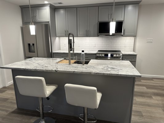 Basement Apartment Remodel - Once an unfinished basement cluttered with exposed utilities and debris, this space has been completely transformed. The owner’s vision to clear mechanical components led to a sleek, modern living area. Luxurious vinyl plank flooring, stainless steel appliances, elegant gray cabinetry, and a marble-patterned island now define the space. Warm pendant lighting completes this stylish and functional retreat.
