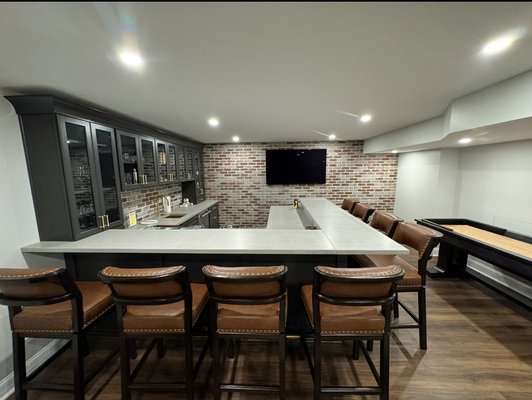 Custom Basement Build Out with Wet Bar, Golf Simulator, Wine Cellar, and Gym - We transformed this unfinished basement into a fully customized lower level designed for entertaining, family time, and everyday functionality. The project began with structural excavation and underpinning to lower the slab and create the proper ceiling height for a dedicated Trackman golf simulator. This phase required careful planning and coordination to safely support the existing foundation while expanding the usable space below.

Once the structural work was complete, we built out a full custom wet bar equipped with a built-in dishwasher, ice machine, and multiple refrigeration units to create a true entertainment hub. The bar features custom poured concrete countertops along with a matching raised concrete bar top that overlooks the golf simulator bay, creating a clean, modern focal point that ties the entire space together.

Adjacent to the bar, we constructed a fully conditioned wine cellar with proper insulation and climate control to ensure long-term preservation. The lower level was designed to serve the entire family, so we added a dedicated home gym, a custom closet for additional storage, and a built-in homework station for the kids featuring integrated cabinetry and a shiplap accent wall for added character. New flooring was installed throughout to create a seamless, cohesive finish across all areas.

This project highlights our ability to manage complex structural modifications alongside detailed finish carpentry and custom fabrication, delivering a thoughtfully designed space built for both performance and long-term durability.