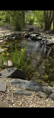 Custom Two-Tier Koi Pond with Waterfall Connection & Aquatic Plant Design - Professionally designed and installed koi pond featuring two separate water sections connected by a natural stone waterfall for continuous circulation and visual appeal. This project included full excavation, liner installation, natural boulder placement, and integration of aquatic plants to create a balanced and low-maintenance ecosystem. Decorative river rock and strategic plant placement enhance the natural appearance while providing a peaceful outdoor environment. This custom water feature adds beauty, movement, and a relaxing focal point to the landscape.