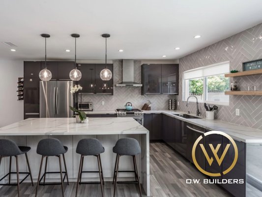 Kitchen, Living Room Remodel | San Francisco - This open-concept kitchen remodel pairs glossy grey cabinetry with a striking quartz waterfall island and sleek herringbone backsplash. The result is a seamless blend of contemporary luxury and functional design, anchored by wood-look flooring and designer lighting throughout.
