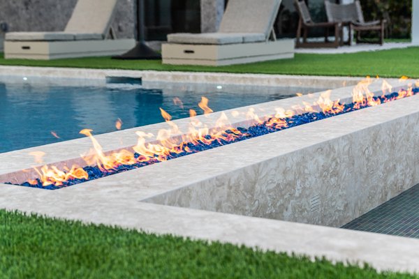 701 S Ocean Project - Imagine a stunning luxury pool set in an expansive outdoor oasis. The pool features sleek, modern design with medium blue water that sparkles under the sun. At one end, a long, linear fire feature runs along the edge, where flames dance on a bed of smooth, dark stones, casting a warm, inviting glow.
The fire feature is integrated seamlessly into the pool's architecture, with the flames seemingly floating above the water's surface, creating a striking contrast.
Soft, ambient lighting around the perimeter highlights the pool’s elegant contours, while the fire's warm hues reflect off the water, adding a magical atmosphere as night falls. Lounge chairs, plush cushions, and strategically placed decor complete the serene, sophisticated setting. Whether you're enjoying a peaceful evening swim or relaxing by the fire, the space is designed to offer both relaxation and visual wonder. 701 S Ocean Project - Imagine a stunning luxury pool set in an expansive outdoor oasis. The pool features sleek, modern design with medium blue water that sparkles under the sun. At one end, a long, linear fire feature runs along the edge, where flames dance on a bed of smooth, dark stones, casting a warm, inviting glow.
The fire feature is integrated seamlessly into the pool's architecture, with the flames seemingly floating above the water's surface, creating a striking contrast.
Soft, ambient lighting around the perimeter highlights the pool’s elegant contours, while the fire's warm hues reflect off the water, adding a magical atmosphere as night falls. Lounge chairs, plush cushions, and strategically placed decor complete the serene, sophisticated setting. Whether you're enjoying a peaceful evening swim or relaxing by the fire, the space is designed to offer both relaxation and visual wonder.