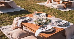 Luxury Picnic Setup - A vibrant blanket on lush grass, surrounded by tall trees and a serene lake. A wicker picnic basket with gourmet sandwiches, fruits, cheese, a bottle of wine, and two glasses chills nearby. Scattered pillows and a designer frisbee invite relaxation and play.