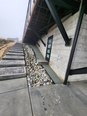 Permeable Paver Steps & Drainage Swale - A property developer working on a new home in Dillon Beach asked us to install some stair treads and a swale. But there were some stipulations from the architects.

1) The steps must be permeable. 
2) There must be walkable area along the foundation. 
3) There must be a way for the gutter water to get carried away from the foundation while keeping as much on the property as possible. 

No problem, my office bound, projector using friends! We have solutions that work good and look good. 

1) Permeable steps. 
The customer wanted black pavers. We couldn't find a black paver supplier that would deliver a small minimum under 1000 square feet, so we settled on mottled grey and called our friends at Dryscape Materials to deliver 2 pallets of Keystone Permeable Pavers, in Chicago blend. Permeable Pavers allow water to pass through and prevent those annoying puddles that splash on your nice shoes. We set them on a sturdy foundation of 6 inches of drain rock so the water would move through freely. 
2) For the walkable area, the client decided on decomposed granite. Getting the compactor in there was quite difficult, but the team figured it out without me. 
3) The Swale was constructed with 6 inch drain rocks on the middle and 3 inch drain rocks on the sides, creating a more natural look without screwing up the architect's specifications. 

Well done, team! This brutalist beauty is ready for safe, surefooted enjoyment in all conditions.