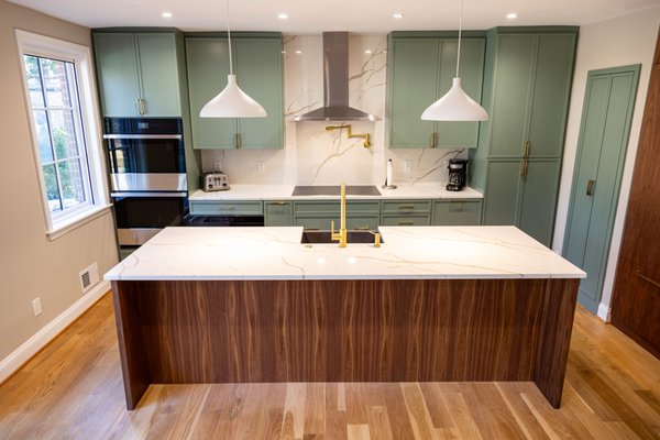 Modern Skinny Shaker Kitchen – Custom Project in Washington, DC - This thoughtfully designed kitchen blends contemporary elegance with timeless craftsmanship. Created for a client in Washington, DC, the space features skinny shaker-style cabinetry in a soft sage green, finished in a satin sheen that adds subtle luster without overpowering the space. Paired with brushed brass hardware, the cabinetry strikes a perfect balance between modern sophistication and classic charm.
The centerpiece is a waterfall-edge walnut island, custom-built to highlight the rich, natural grain of the wood. Topped with a white quartz countertop with elegant veining, the island is both a statement piece and a practical workspace, complete with an integrated sink and satin brass faucet for a cohesive design.
Additional highlights include:
Integrated appliances such as double wall ovens and a paneled refrigerator for a seamless look.
Book-matched marble backsplash for a clean, continuous visual line behind the cooktop.
Contemporary pendant lighting that complements the brass accents and creates warm, focused light.
Floor-to-ceiling cabinetry and a custom pantry that provide both function and visual continuity.
This project is a true example of refined craftsmanship, where satin-finished surfaces, warm walnut, and clean lines come together to create a kitchen that’s as functional as it is beautiful. Modern Skinny Shaker Kitchen – Custom Project in Washington, DC - This thoughtfully designed kitchen blends contemporary elegance with timeless craftsmanship. Created for a client in Washington, DC, the space features skinny shaker-style cabinetry in a soft sage green, finished in a satin sheen that adds subtle luster without overpowering the space. Paired with brushed brass hardware, the cabinetry strikes a perfect balance between modern sophistication and classic charm.
The centerpiece is a waterfall-edge walnut island, custom-built to highlight the rich, natural grain of the wood. Topped with a white quartz countertop with elegant veining, the island is both a statement piece and a practical workspace, complete with an integrated sink and satin brass faucet for a cohesive design.
Additional highlights include:
Integrated appliances such as double wall ovens and a paneled refrigerator for a seamless look.
Book-matched marble backsplash for a clean, continuous visual line behind the cooktop.
Contemporary pendant lighting that complements the brass accents and creates warm, focused light.
Floor-to-ceiling cabinetry and a custom pantry that provide both function and visual continuity.
This project is a true example of refined craftsmanship, where satin-finished surfaces, warm walnut, and clean lines come together to create a kitchen that’s as functional as it is beautiful.
