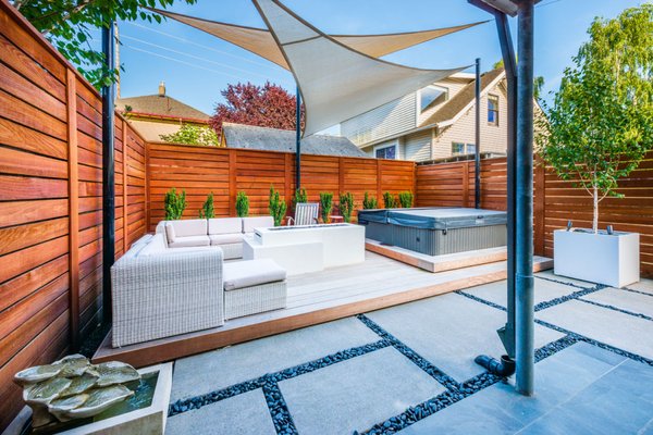 Modern Spa Oasis - This backyard spa retreat’s concrete pad with black polished stones really leans into the contemporary look, while the wood deck and fence make the space warm and inviting. Overhead, the white and cream-colored shade sails not only look great but they also provide privacy from neighboring windows and help the backyard feel more like a room than a fishbowl. The highlights of the yard of course are the hot tub and custom-built fireplace, which includes a shelf to set your drinks on (the client’s own idea!). Low-maintenance evergreen shrubs are planted in a row behind the deck and a large, white, cube-shaped pot has a Himalayan Birch tree planted in it with up lights to help it stand out at night. Modern Spa Oasis - This backyard spa retreat’s concrete pad with black polished stones really leans into the contemporary look, while the wood deck and fence make the space warm and inviting. Overhead, the white and cream-colored shade sails not only look great but they also provide privacy from neighboring windows and help the backyard feel more like a room than a fishbowl. The highlights of the yard of course are the hot tub and custom-built fireplace, which includes a shelf to set your drinks on (the client’s own idea!). Low-maintenance evergreen shrubs are planted in a row behind the deck and a large, white, cube-shaped pot has a Himalayan Birch tree planted in it with up lights to help it stand out at night.