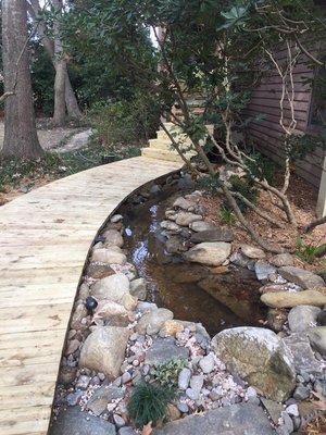 Curved wooden deck walkway over water feature - We renovated the entryway to this beautiful home in Charleston, SC. The entrance access to the front door was a cracked concrete walkway, which we demoed ￼and constructed this beautifully curving elevated wooden walkway over a water feature that we also installed . When the owner sold the home they got an offer for $30,000 over the asking price specifically because of the work they had done in the landscaping, principally this walkaway with the water feature that we installed. 
.