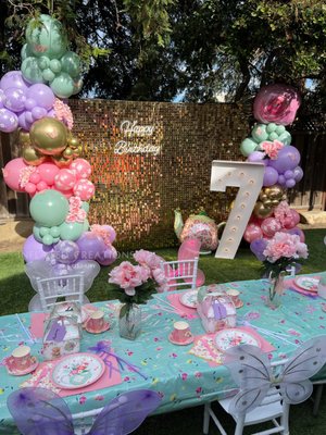 7th Birthday Par-tea - This styling was done for a 7 year old birthday party. She wanted a fun tea party with flowers, pastels and all things girly! We did a custom acrylic welcome sign along our copper frame, adorned with balloons, butterflies and flower accents. This greeted guests as they arrived. We then created a fun table scape with flower vases, tea party plates, cups and cutlery, added a fairy wand and custom party favor boxes and dressed up the chairs with fairy wings. We then installed a beautiful gold shimmer wall backdrop with an organic balloon garland, butterfly and flower accents, specialty foil balloons, light up marquee and neon Happy Birthday sign.