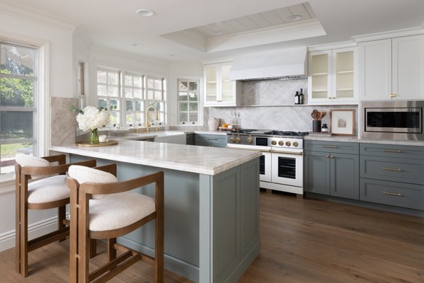 Bright, Timeless Kitchen Remodel in San Juan Capistrano - A well-designed kitchen should support the way you live while reflecting your personal style. This 400-square-foot kitchen remodel in San Juan Capistrano transforms a dated layout into an inviting space that blends classic design with modern functionality. Soft finishes, natural materials, and custom craftsmanship come together to create a kitchen that's equally suited for entertaining and everyday living.

An Open Layout That Invites Connection
At the center of the design is a spacious peninsula with seating for two, creating a casual spot for meals, conversation, or helping with dinner preparation. The expanded work surface improves functionality while maintaining an open, comfortable flow throughout the kitchen.
Wide-plank European oak engineered hardwood flooring in the color Verte anchors the space with rich natural texture and complements the kitchen's warm, neutral palette.

Custom Cabinetry with Distinctive Character
Kemper maple cabinetry provides extensive storage while introducing subtle contrast through a two-tone finish. The upper cabinets, pantry, paneled refrigerator and freezer are finished in ‘Painted Coconut’, creating a crisp backdrop that keeps the room feeling open and airy.
Below, the base cabinetry is finished in ‘Painted Retreat’, adding depth with a soft green hue that pairs beautifully with the surrounding materials.

Four upper cabinets feature reeded glass inserts, offering visual interest and an elegant place to display glassware and decorative pieces. A paneled dishwasher disappears into the cabinetry, preserving the clean, uninterrupted lines of the design.
Throughout the kitchen, aged brass cabinet knobs and appliance pulls introduce warmth and tie the finishes together with understated elegance.

Natural Stone That Makes a Statement
The countertops and full-height backsplash are crafted from stunning Taj Mahal quartzite, a natural stone prized for its soft veining, exceptional durability, and timeless beauty.
Extending the quartzite from the counter-top up the backsplash creates a seamless, luxurious backdrop while allowing the cabinetry and brass accents to remain the focal points.

A Chef-Inspired Cooking Space
The cooking area is designed for both performance and style, featuring a 48-inch slide-in range paired with a built-in microwave for convenience.
Above the range, a custom sloped hood finished in La Llabra Crystal White Smooth micro-cement serves as a striking architectural feature. Its clean silhouette adds subtle texture while complementing the surrounding cabinetry and natural stone.

Functional Finishes with Lasting Appeal
Positioned beneath expansive windows, the Kohler Whitehaven undermount single-bowl farmhouse sink in Sea Salt White provides generous workspace and takes full advantage of the natural light.
A satin gold high-spout squeeze-handle faucet and coordinating soap dispenser echo the aged brass hardware, bringing cohesion to the overall design.

Designed for Everyday Living
Flooded with natural light, this newly remodeled kitchen showcases a fresh, timeless palette featuring soft white cabinetry, muted green lower cabinets, warm aged brass accents, and luxurious Taj Mahal quartzite surfaces. Every detail was carefully selected to maximize storage, improve workflow, and create a welcoming atmosphere for cooking, entertaining, and family life.
The finished result is a timeless, light-filled space that will serve as the heart of the home for years to come.

Contact Us
If you’re planning your own Kitchen remodel in San Juan Capistrano or any other home remodeling project in Orange County, Sea Pointe Design & Remodel has you covered! We’ve been meeting the remodeling needs of Orange County residents for over 35 years, and we’d love to remodel YOUR home next. Simply give us a call at (949) 861-3400 or fill out the form below to schedule your complimentary design consultation.