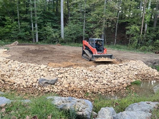 Fire pit build! - This customer wanted a fire pit hangout on the other side of a stream that flowed through his property, we built a temporary bridge to get our machine across to grade, add river rock for erosion control, landscape fabric and metal edging to separate the rock from the mulch we lined on the outside of the fire pit. The customer was super happy with the result and so were we!