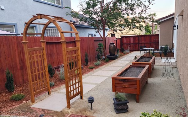 Garden-Focused Design with Clean, Functional Layout - This project was designed to create a simple, organized backyard with a focus on usability and a dedicated gardening space. We installed a decomposed granite patio and pathway with concrete steppers to define movement through the yard and create a clean, structured layout. A custom wood arbor adds a natural focal point and entry into the space, while raised planter beds were built to provide a functional and accessible gardening area.
The perimeter was finished with low-maintenance planting, fresh mulch, and subtle landscape lighting to keep the space clean and easy to manage. A decorative water feature adds a calming element and helps tie the space together.The result is a practical, low-maintenance backyard that blends gardening, relaxation, and clean design. Garden-Focused Design with Clean, Functional Layout - This project was designed to create a simple, organized backyard with a focus on usability and a dedicated gardening space. We installed a decomposed granite patio and pathway with concrete steppers to define movement through the yard and create a clean, structured layout. A custom wood arbor adds a natural focal point and entry into the space, while raised planter beds were built to provide a functional and accessible gardening area.
The perimeter was finished with low-maintenance planting, fresh mulch, and subtle landscape lighting to keep the space clean and easy to manage. A decorative water feature adds a calming element and helps tie the space together.The result is a practical, low-maintenance backyard that blends gardening, relaxation, and clean design.