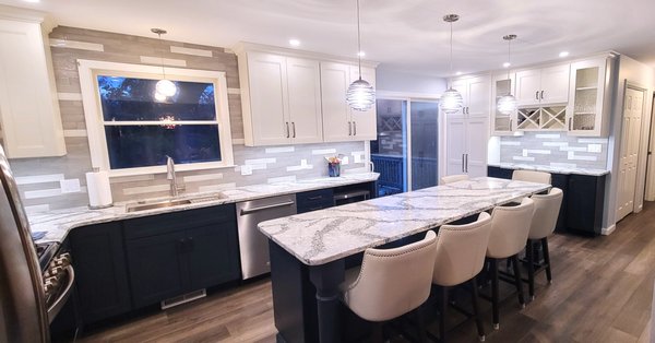 Modern Elegance with Expansive Granite Island - This kitchen remodel project beautifully combines modern elegance with functionality. The highlight of the space is the expansive granite island, featuring intricate veining and a polished finish that adds a touch of luxury. Surrounding the island are custom-built white cabinetry that offers ample storage and seamlessly blends with the gray and navy lower cabinets, creating a striking contrast. The backsplash, composed of horizontal tiles in varying shades of gray, complements the stainless steel appliances and the sleek hood, providing a cohesive look. Pendant lights above the island add a sophisticated touch, while the under-cabinet lighting enhances the workspace. This remodel not only enhances the aesthetic appeal of the kitchen but also improves its functionality, making it the perfect space for both cooking and entertaining. Modern Elegance with Expansive Granite Island - This kitchen remodel project beautifully combines modern elegance with functionality. The highlight of the space is the expansive granite island, featuring intricate veining and a polished finish that adds a touch of luxury. Surrounding the island are custom-built white cabinetry that offers ample storage and seamlessly blends with the gray and navy lower cabinets, creating a striking contrast. The backsplash, composed of horizontal tiles in varying shades of gray, complements the stainless steel appliances and the sleek hood, providing a cohesive look. Pendant lights above the island add a sophisticated touch, while the under-cabinet lighting enhances the workspace. This remodel not only enhances the aesthetic appeal of the kitchen but also improves its functionality, making it the perfect space for both cooking and entertaining.