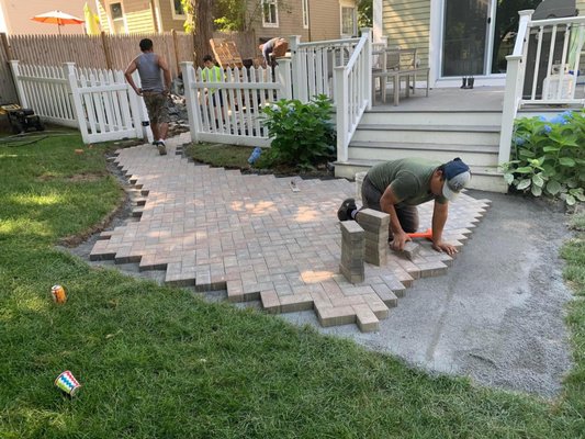 Herringbone Patio w dark border - The backdoor to the driveway was a heavily-trafficked but overlooked social area. The homeowners not only created a patio for entertaining off the kitchen, but solved a major headache with a graceful walkway with access to driveway (think groceries.)

Over time, landscaping beyond the grey-stone border, and furniture etc., will feature the masonry as the bones and beginning of a well-loved outdoor room.