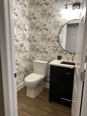 Fun and Flirty Powder Room - High contrast vanity with Thunder Grey Kohler sink with Champagne Bronze Delta Stryke Fixtures wtih skirted toilet, wood tile plank floor with wallpaper finish.