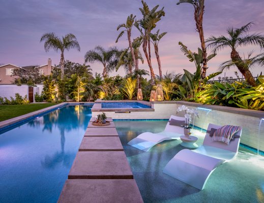 North County Coastal - This client had visions of a Hawaiian vacation resort/oasis for their coastal north county home which needed an update. They selected Team G7 to make their vision a reality. This elegant project includes a contemporary lap pool with a 45-foot-long swim lane, an 8-foot-deep dive area, and a relaxing Baja shelf for adults which also serves as a protected play area for their young grandchildren. This unique design includes “floating” steppers leading to the spa that complement the modern lines of the project while acting as a preventive Baja shelf guard for young children.
The pool connects seamlessly to the cabana which is a brilliant white patio structure with a beautiful Ipe hardwood ceiling, providing an amazing lounge area that includes a large recessed TV and a walk-up kitchen/BBQ area. Adjustable LED lighting is integrated into the landscaping and cabana for “warm” accent lighting and functional use. At night, both the pool and the sheer descent waterfall colors can be controlled remotely, contributing to an incredible light show enjoyed from the solid patio structure and the home’s windows facing the backyard.
Landscaping was carefully selected to create an oasis feeling that frames and complements the hardscape design. The beautiful layers of green foliage are accented by a “specimen” plumeria tree where the fragrance cannot help but remind you of the Hawaiian islands and bring peace and tranquility to your state of mind. North County Coastal - This client had visions of a Hawaiian vacation resort/oasis for their coastal north county home which needed an update. They selected Team G7 to make their vision a reality. This elegant project includes a contemporary lap pool with a 45-foot-long swim lane, an 8-foot-deep dive area, and a relaxing Baja shelf for adults which also serves as a protected play area for their young grandchildren. This unique design includes “floating” steppers leading to the spa that complement the modern lines of the project while acting as a preventive Baja shelf guard for young children.
The pool connects seamlessly to the cabana which is a brilliant white patio structure with a beautiful Ipe hardwood ceiling, providing an amazing lounge area that includes a large recessed TV and a walk-up kitchen/BBQ area. Adjustable LED lighting is integrated into the landscaping and cabana for “warm” accent lighting and functional use. At night, both the pool and the sheer descent waterfall colors can be controlled remotely, contributing to an incredible light show enjoyed from the solid patio structure and the home’s windows facing the backyard.
Landscaping was carefully selected to create an oasis feeling that frames and complements the hardscape design. The beautiful layers of green foliage are accented by a “specimen” plumeria tree where the fragrance cannot help but remind you of the Hawaiian islands and bring peace and tranquility to your state of mind.